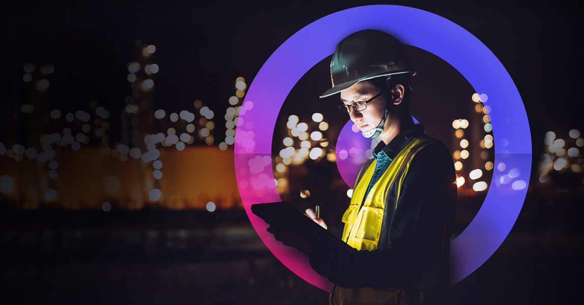 A male engineer wearing a white hard hat and a yellow high vis working on his ipad with lights in the background and a cora lens circling him