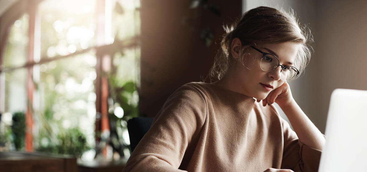 A blonde lady wearing glasses and a brown jumper working on her laptop
