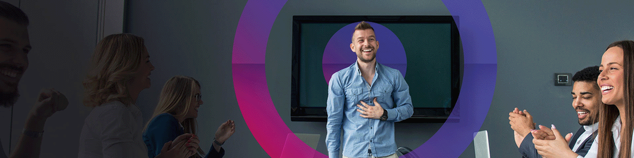 People in an office laughing while discussing work. Man in blue shirt standing in front of a television screen with his hand on chest. Pink and blue Cora lens in the middle centre of image.