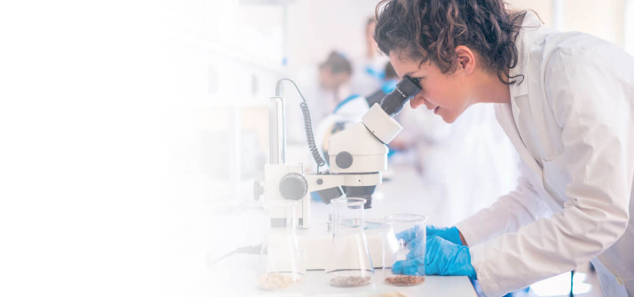 A lady scientist looking through a telescope in a lab wearing blue gloves and a white lab coat. Three glass cylinders beside her.