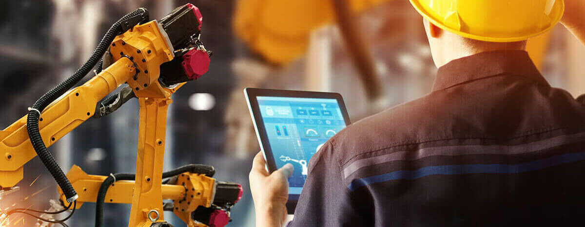 Male engineer wearing a yellow hard hat looking at an ipad in a factory with a yellow robotic machine in the background