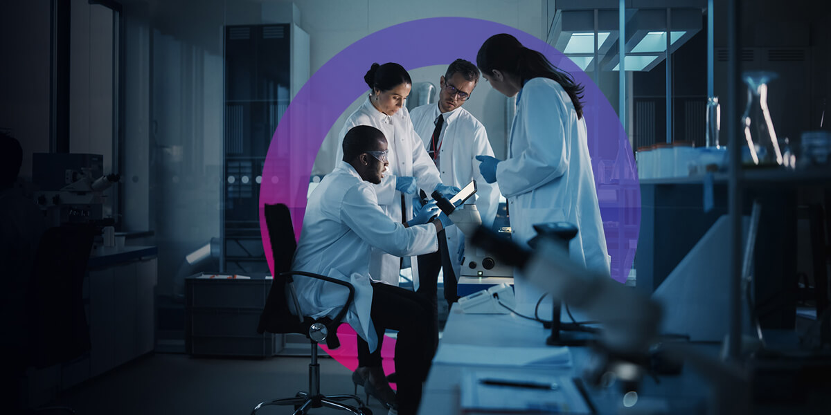 Team of lab analysts in having a team discussion. All of them are wearing white lab coats and safety goggles.