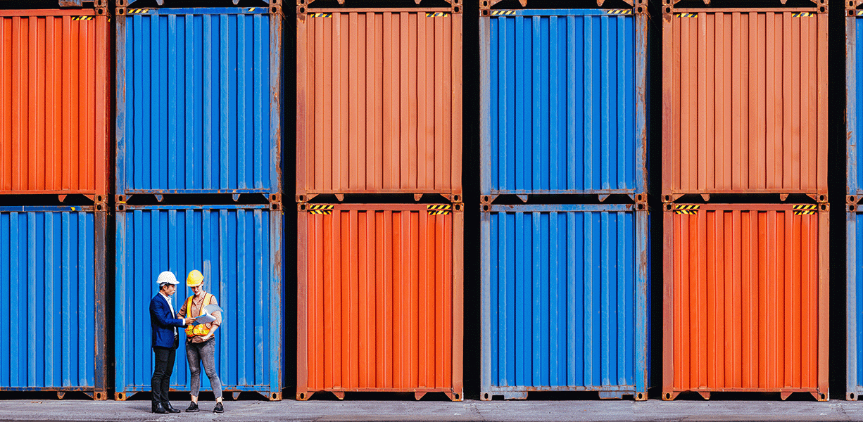 An image of many blue and red storage containers with two engineers standing in front wearing white hard hats and one is wearing an orange high vis 