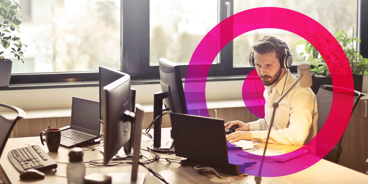 Man wearing headset in an office working at his desk. He is typing on his laptop. Pink and purple gradient lens in the background.