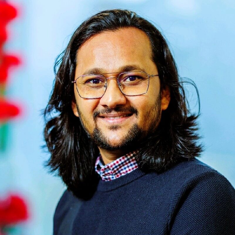 Image of man wearing glasses with long dark hair wearing a navy jumper and shirt.
