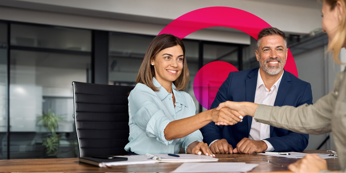 Lady in blue shirt shaking hands with another lady who is out of the frame. Office setting. Pink Cora lens behind lady in blue shirt and man in a navy suit.